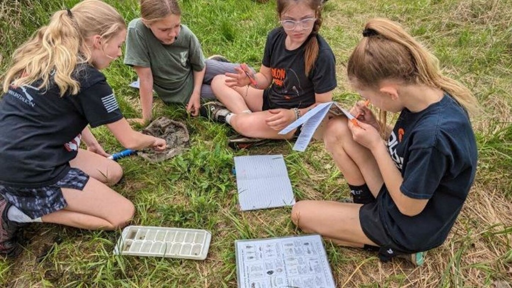 kids outside sitting on the ground, recording their findings during School of the Wild